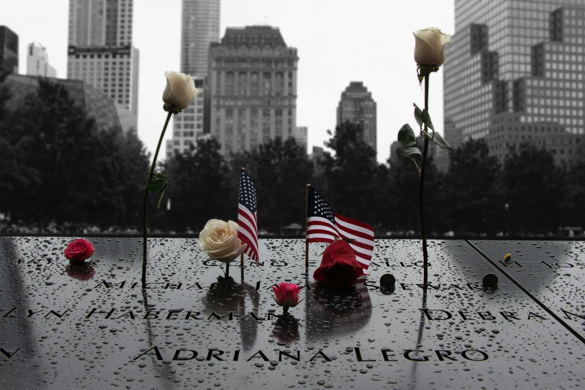 A group of flags on the 9/11 memorial with flowers. Victims name are etched on the memorial.