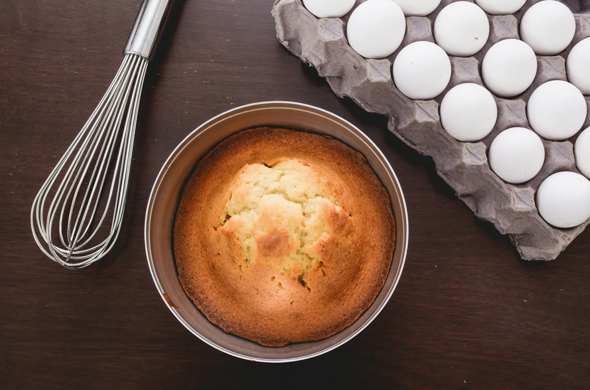 Sponge cake in baking pan with eggs and a whisk kept beside it.