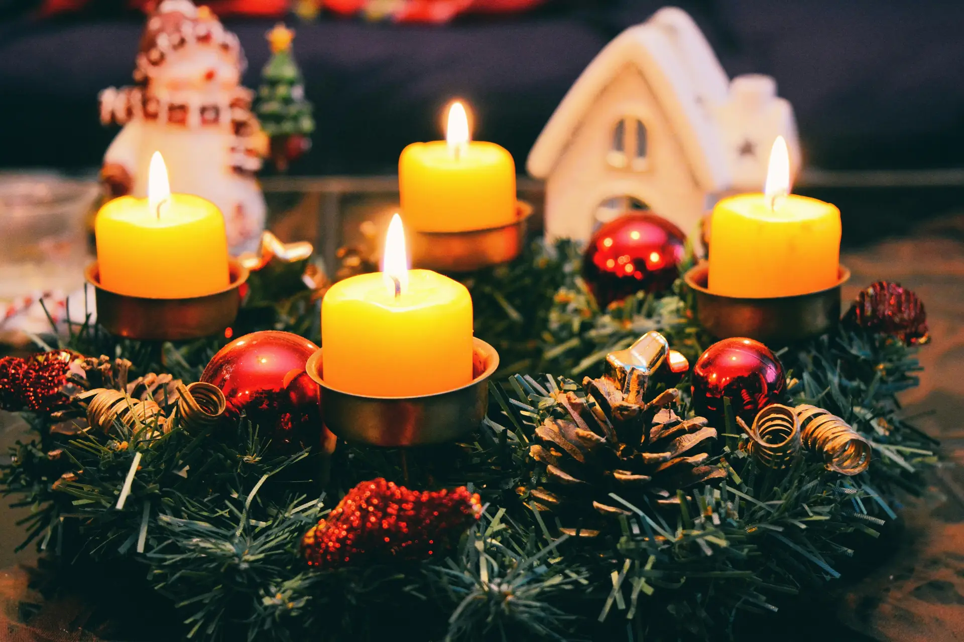 Bunch of candles are lit on a table. There are spruce needles and gingerbread house near them.