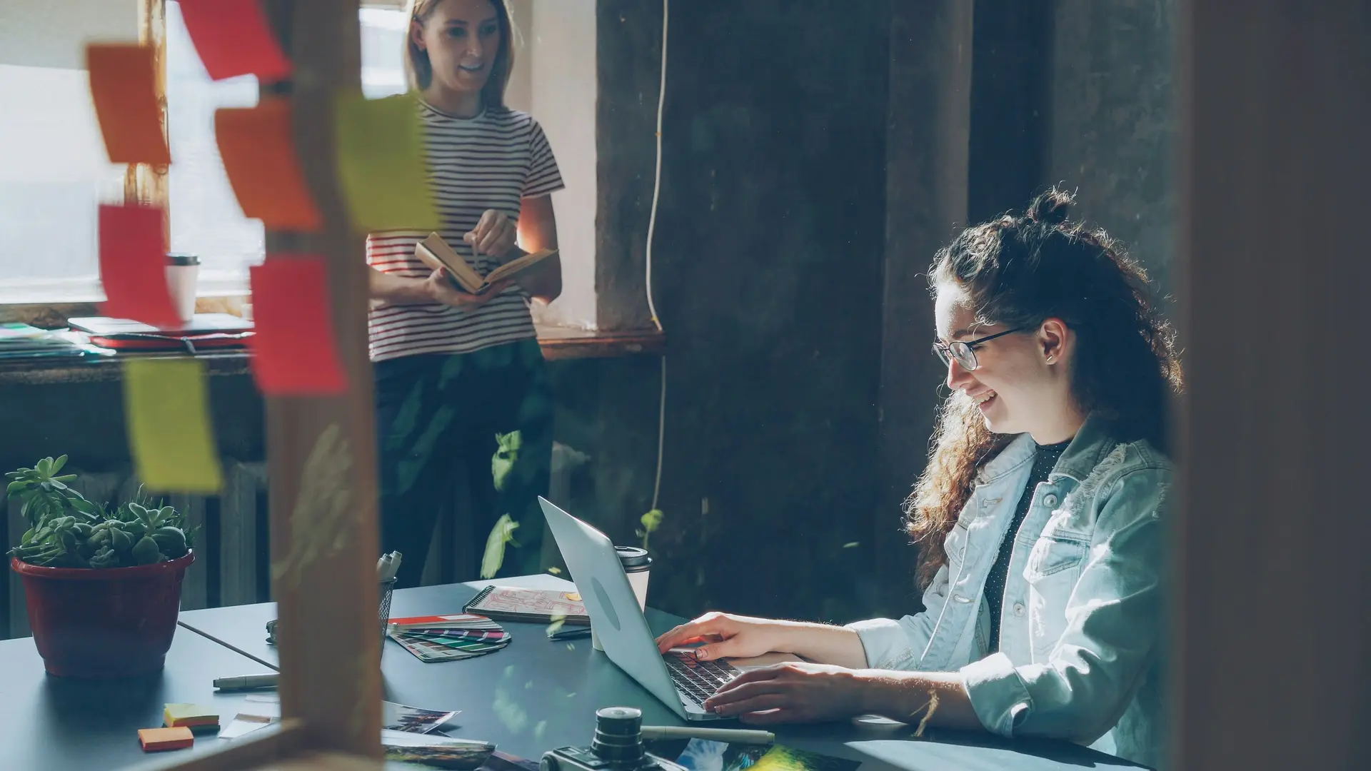 A woman IT expert working on the laptop while another woman standing near her holding a book.
