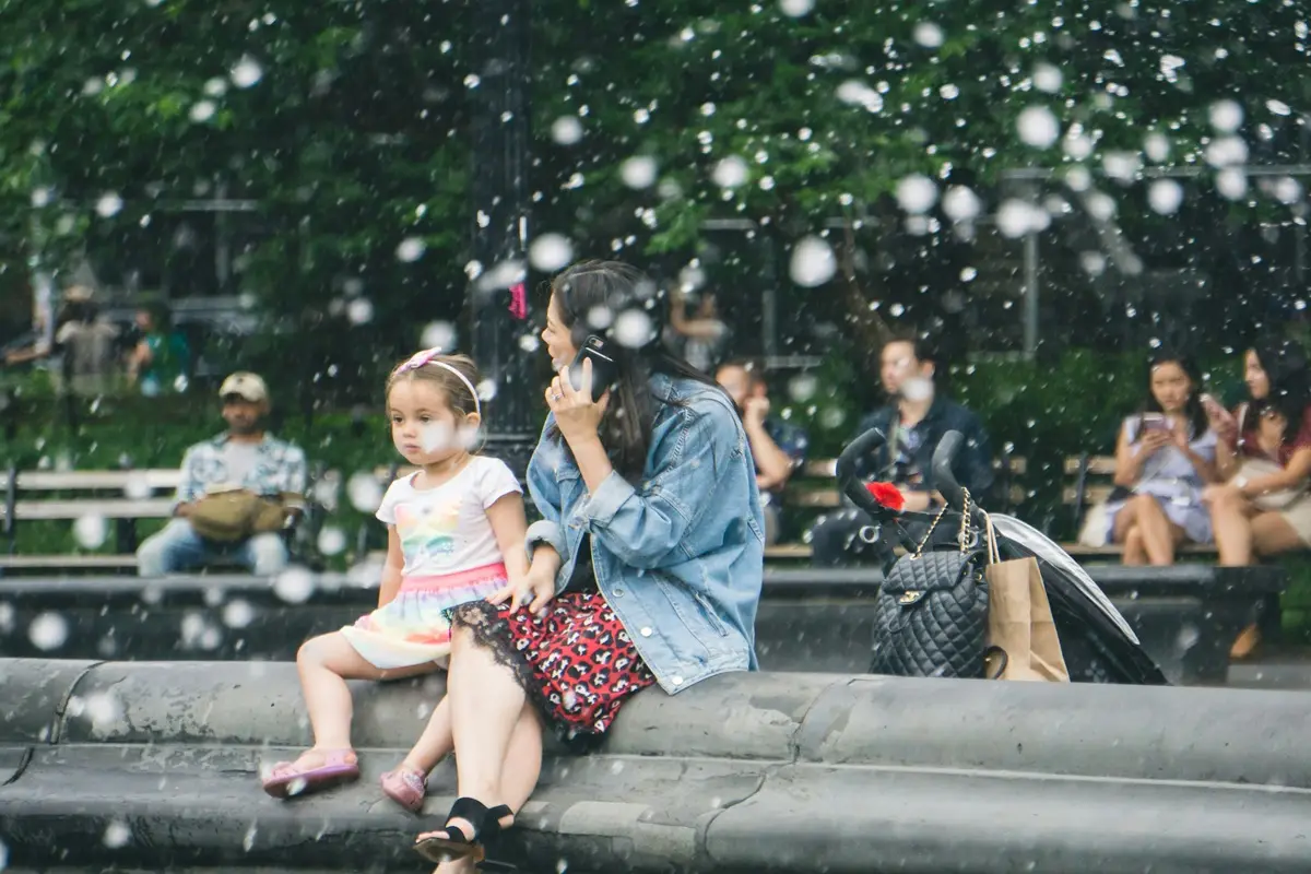 A kid is sitting with her aunt in a park while her aunt is talking over the phone.
