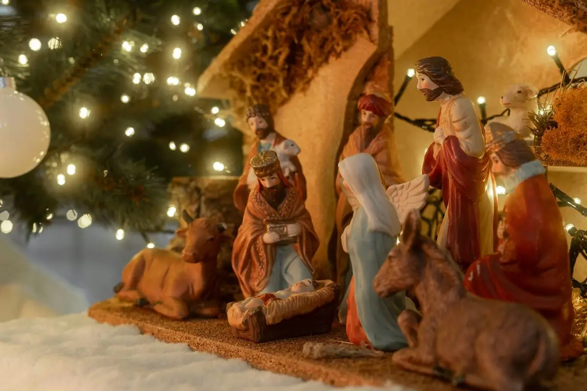 A lit nativity scene made with figurines of Jesus, Mary, Joseph, The Three Kings, and horses. A lit Christmas tree is seen in the background.