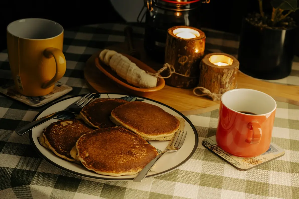 Warm breakfast scene with pancakes, coffee, and candles on a checkered tablecloth.