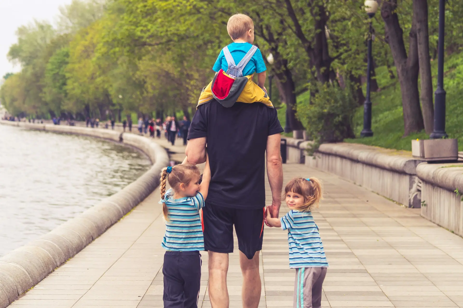 A dad walking with three of his children. One is one the shoulder and other two is grabbing his two hands.