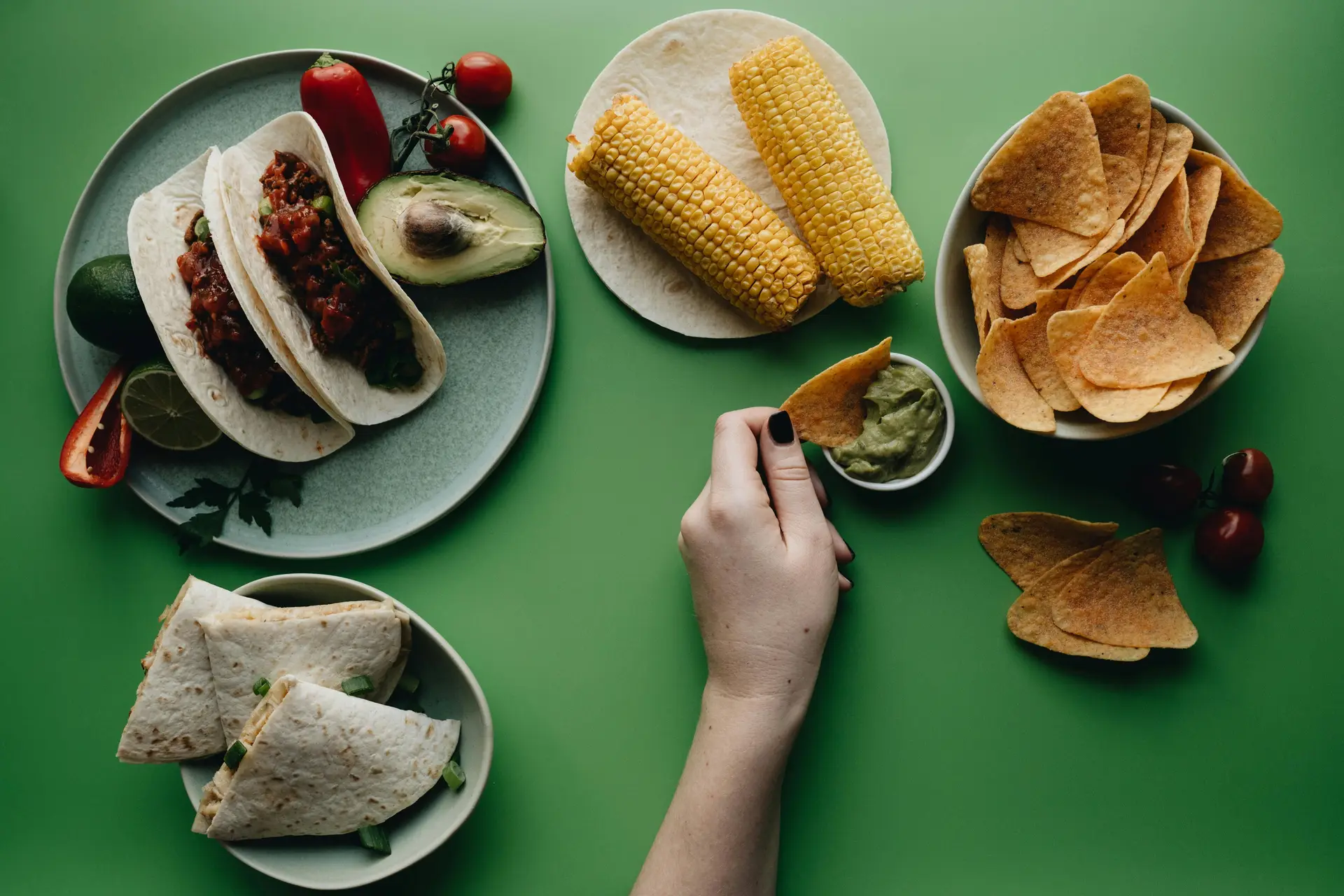 A person's hand seen dipping a chip in guacamole. There are corn, more chips and other food items on the table along with the dip.