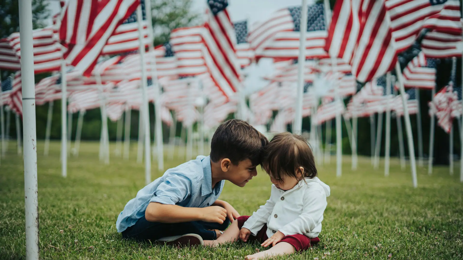 Little brother and sister sitting on the grass and there are a lots of usa flags arond them.