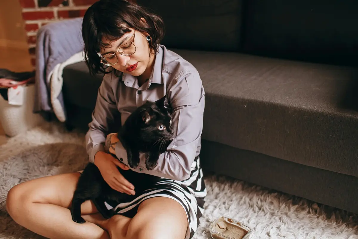 Woman holding a black cat while sitting on a cozy rug indoors.