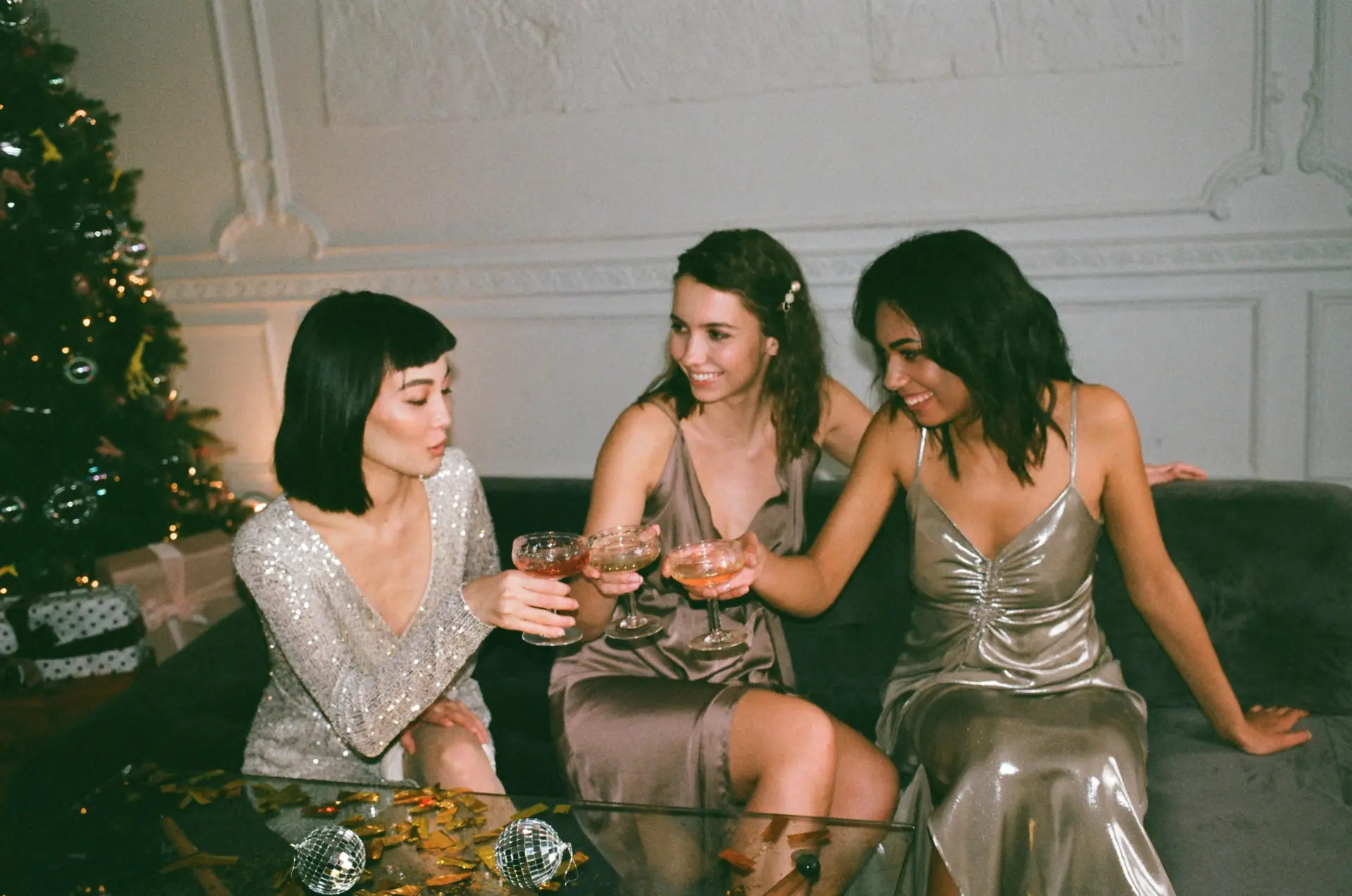 Three women sitting on a sofa and enjoying drinks. They are wearing party dresses.