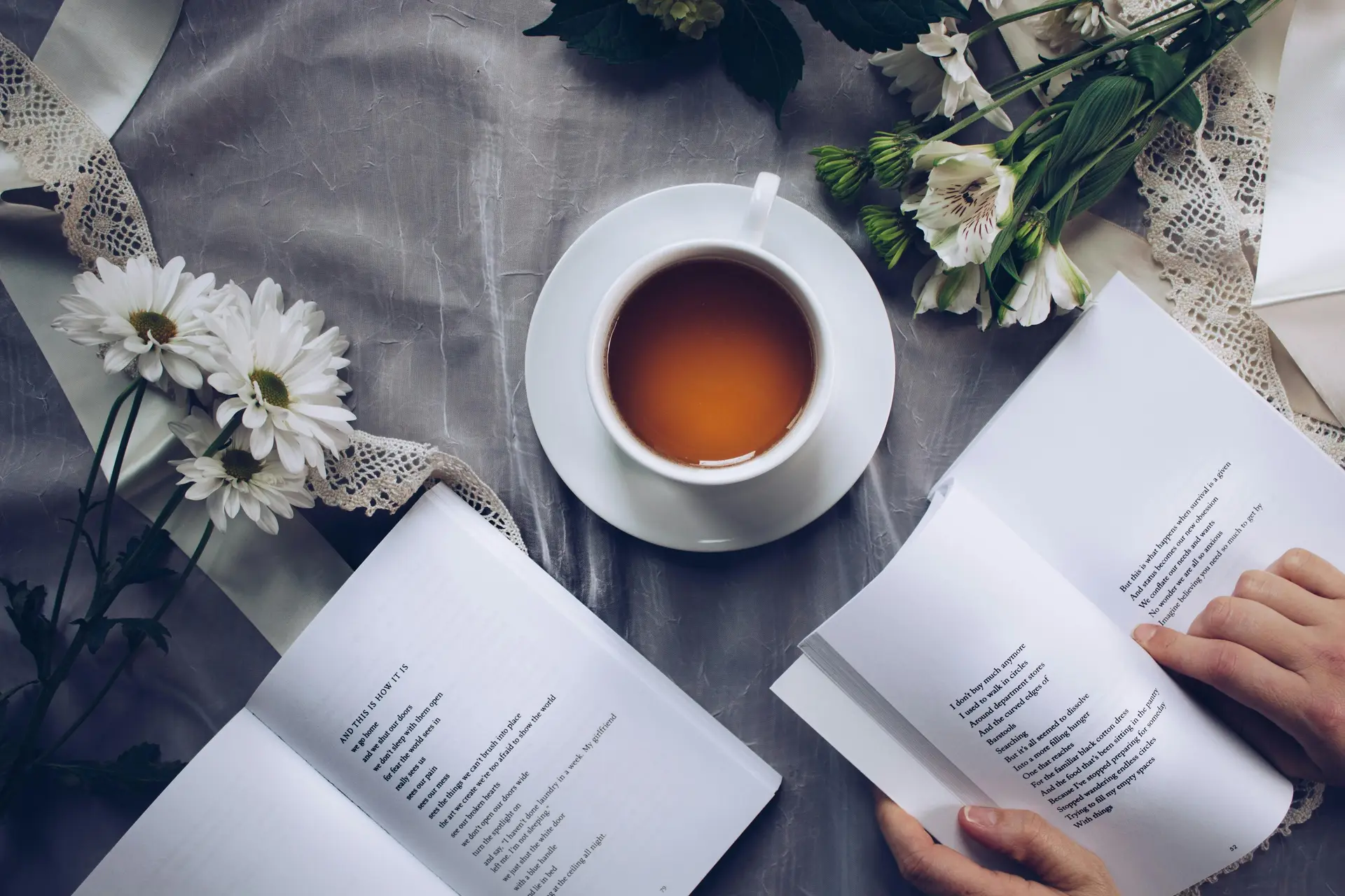 A cup of coffee placed on a gray floral textile cover along with two open poetry books and bunch of flowers.