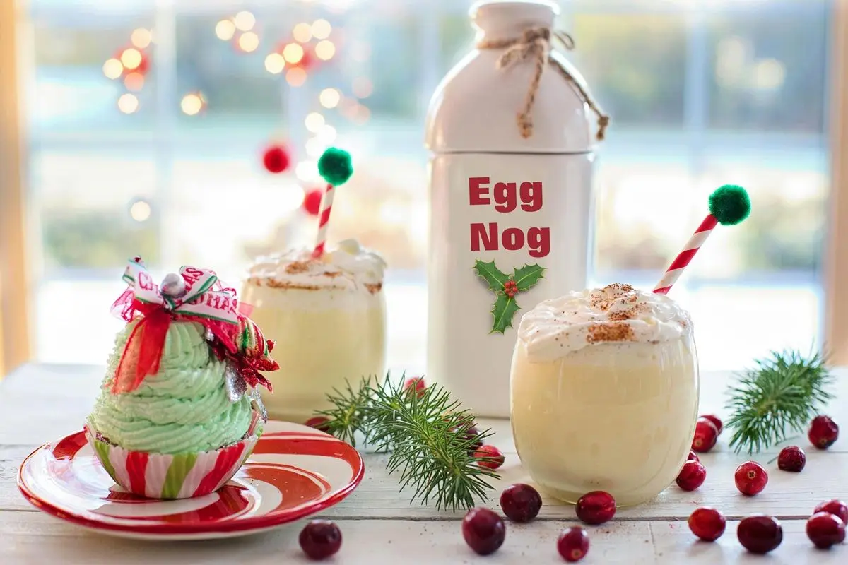 On a table there's a eggnog filled bottle with  Egg Nog label. There are some cups and cherries on the table as well.
