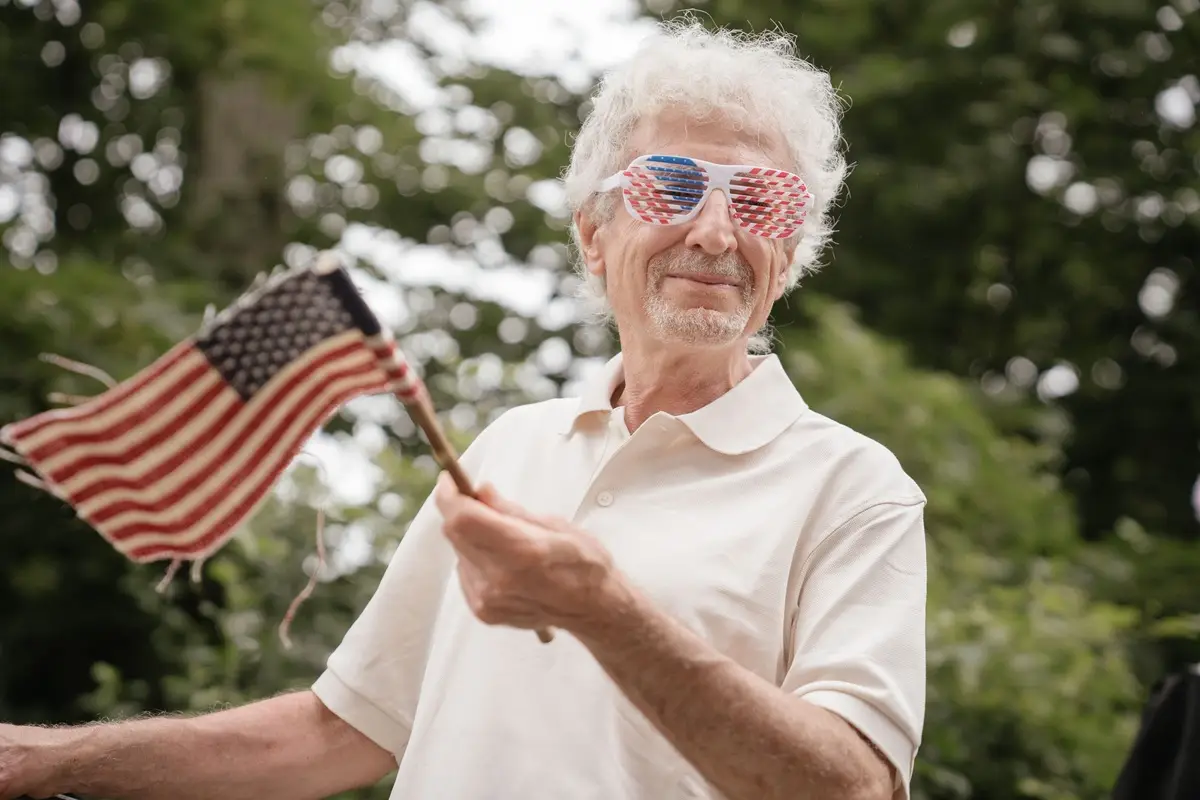 Elderly man with star-spangled sunglasses holding an American flag outdoors.
