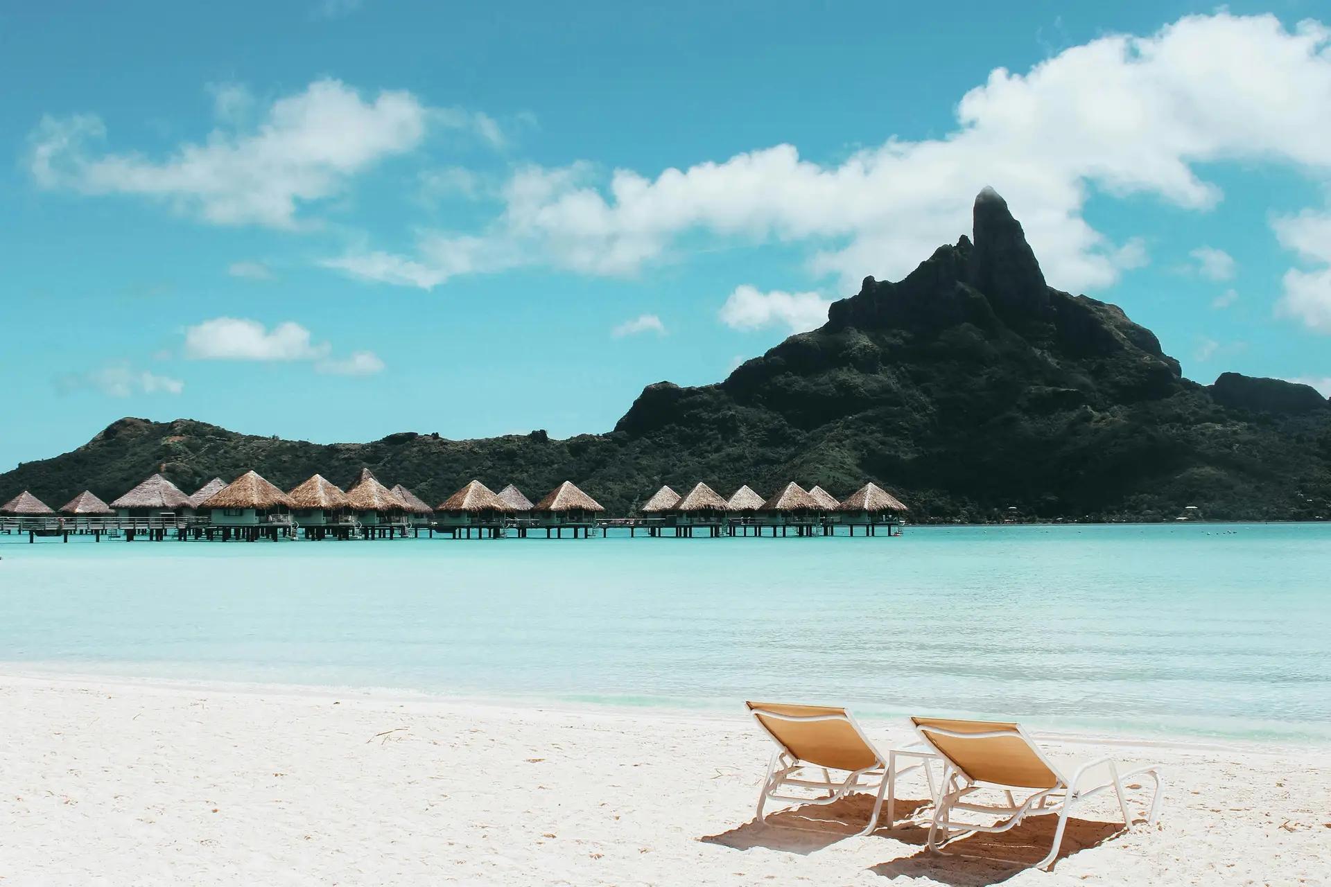 On a beach there are two beach chair along. A mountain and the sea can be seen as well.