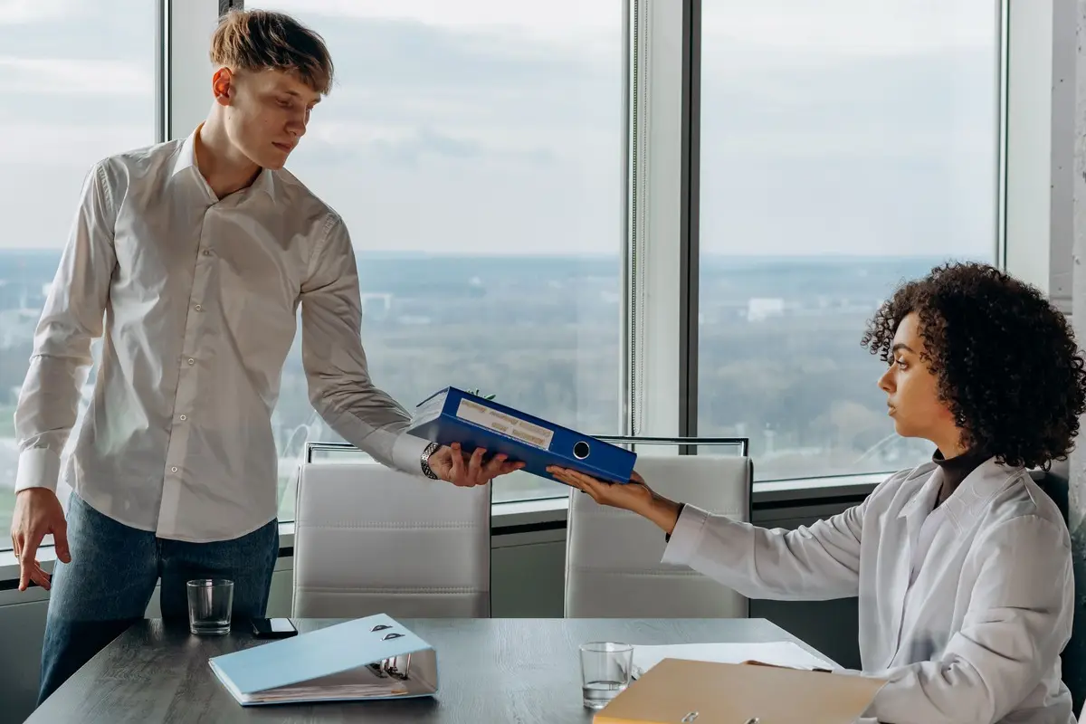 Two colleagues exchange documents in a contemporary office with a scenic city view.