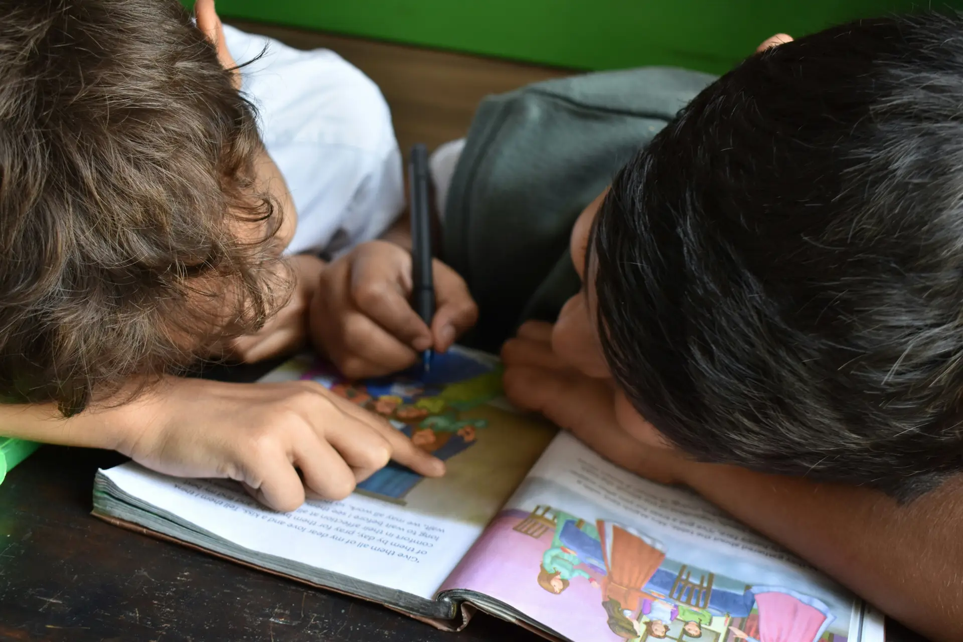 Two kids writing on a book with a black pen.