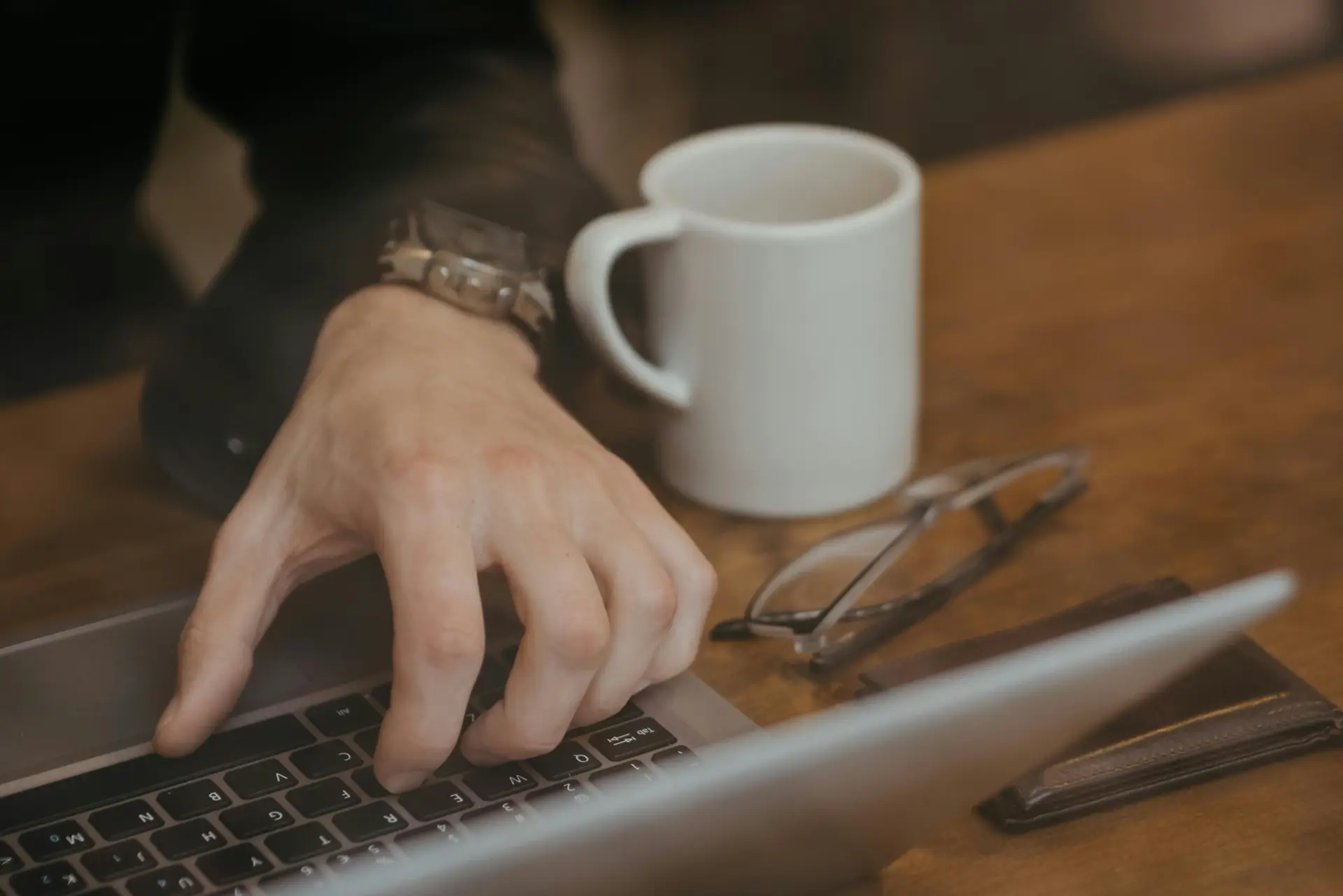 A person using laptop with their left hand. There's a glass and mug on the table along with laptop.
