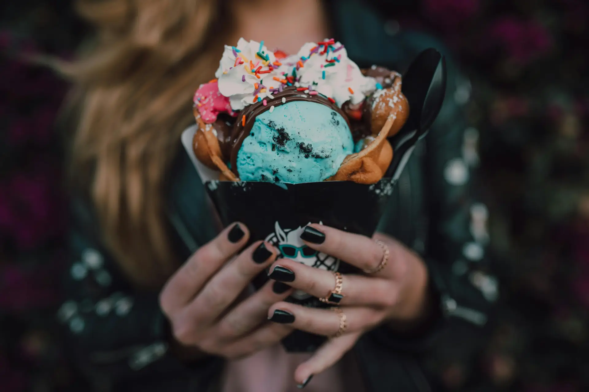 Different flavored ice cream scoops, wafer, syrup and sprinkles used to make a extravagant cup of ice cream which is held by a lady.