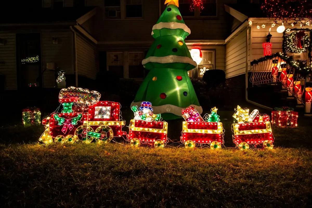 A group of colorful lit train Christmas decoration along with a tree is placed outside a home during nighttime.