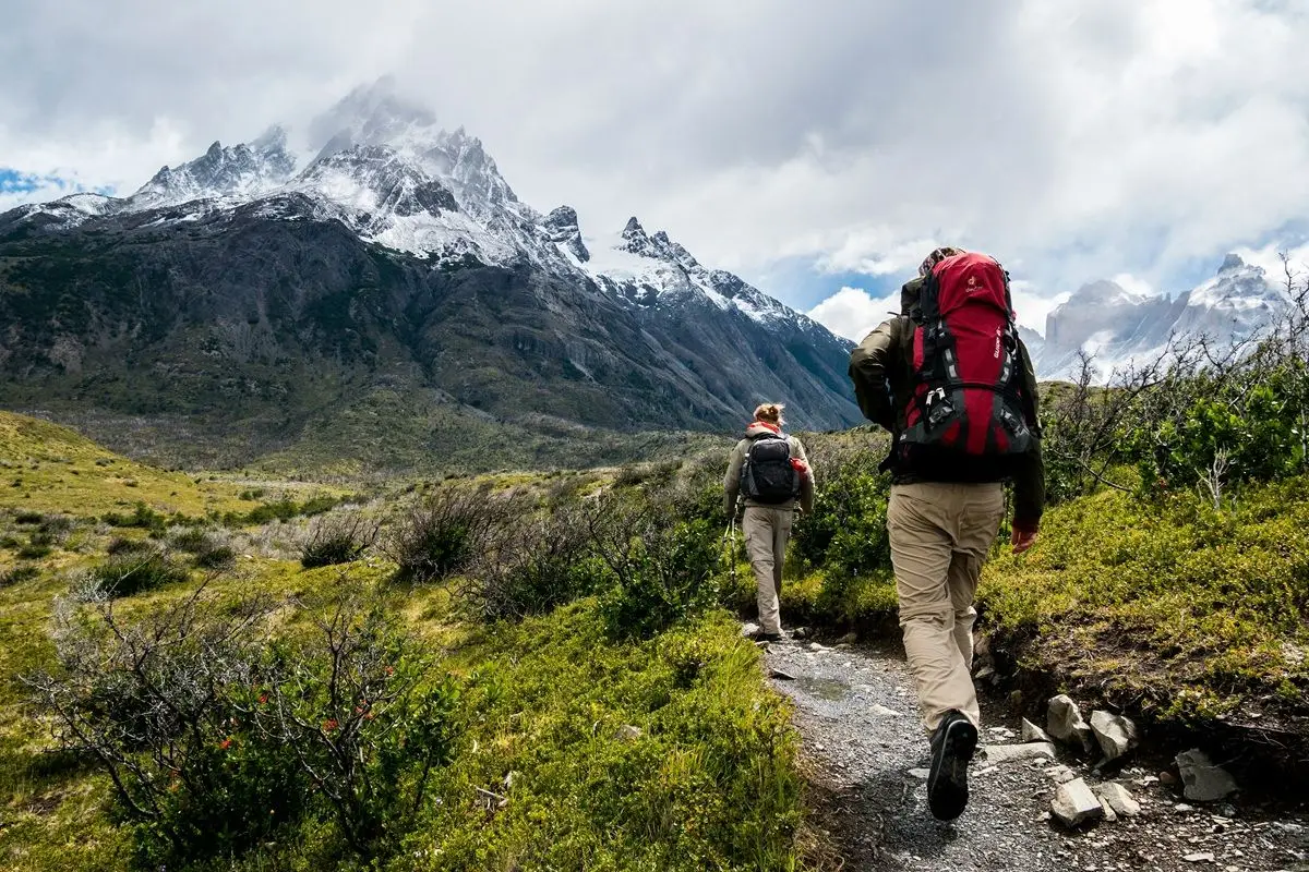 A person with a red backpack is hiking in one of the national park of America with the icy peaks of a mountain can be seen in the distance.