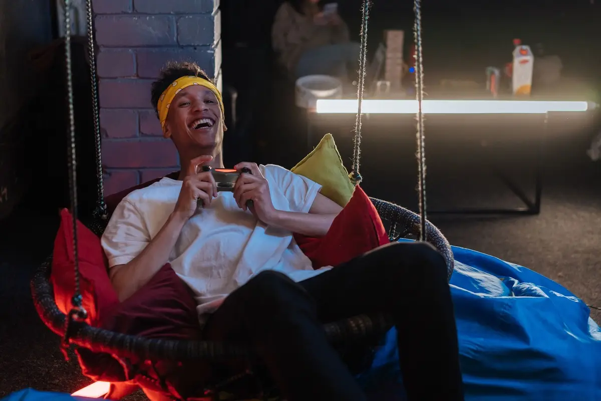 A young man with a joyful expression gaming indoors while sitting on a swing chair.