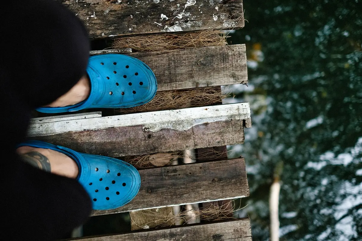 A person in blue crocs standing on a rustic wooden pier above water.