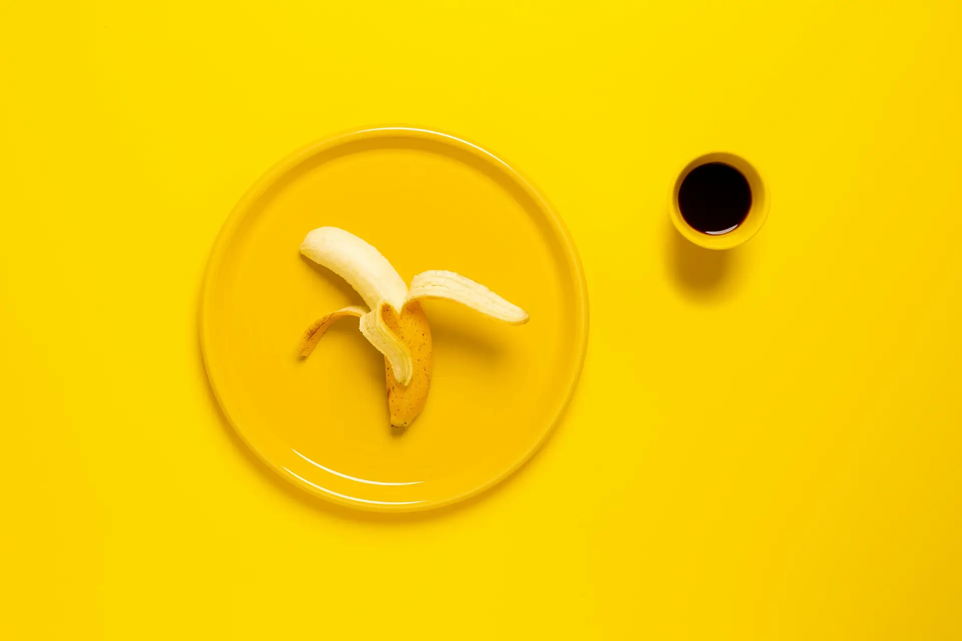 A half peeled yellow banana on a yellow plate, next to a cup of coffee.