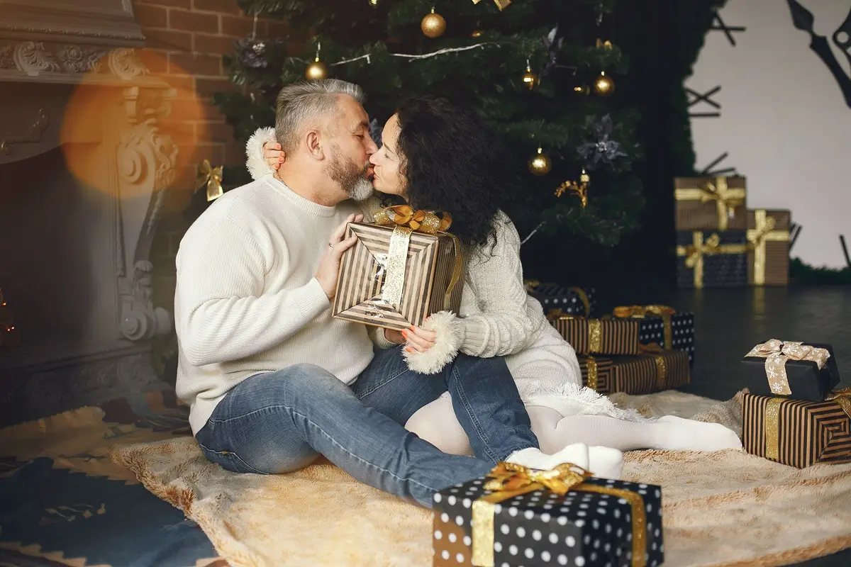A loving couple enjoying a cozy Christmas, exchanging gifts by the Christmas tree, while kissing.