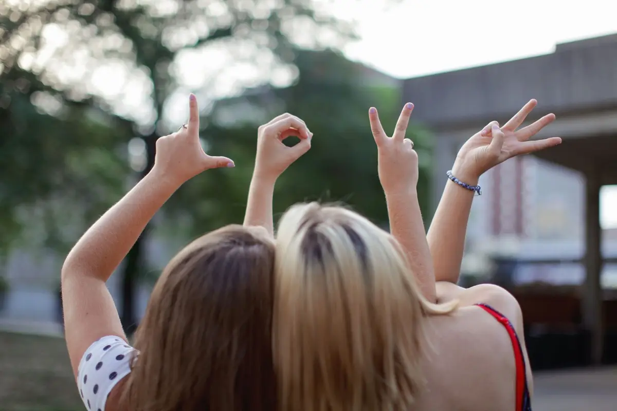 Two young women forming the word 'LOVE' with hand signs, symbolizing friendship in a summer setting.