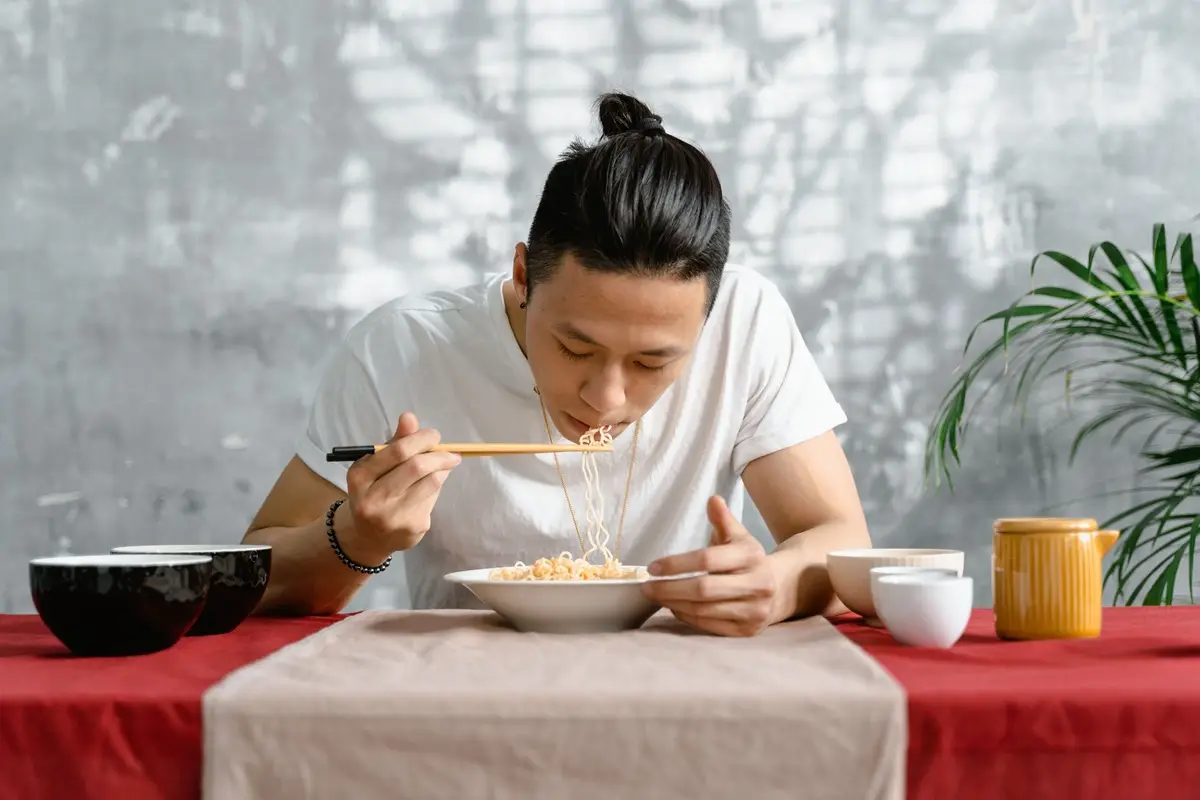 Man in white shirt eating noodles with chopsticks at a table indoors.