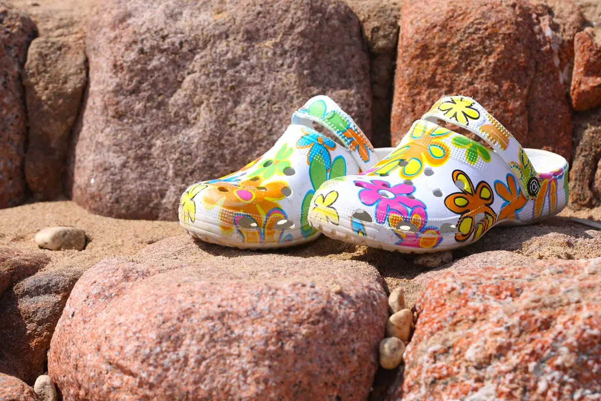 Vibrant floral-patterned clogs displayed on a sandy, rocky beach.
