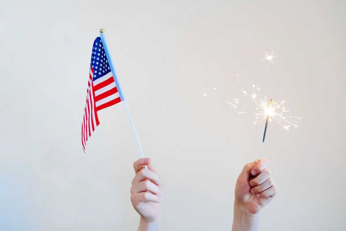 Hands holding an American flag and a sparkler against a white background, symbolizing celebration and patriotism.