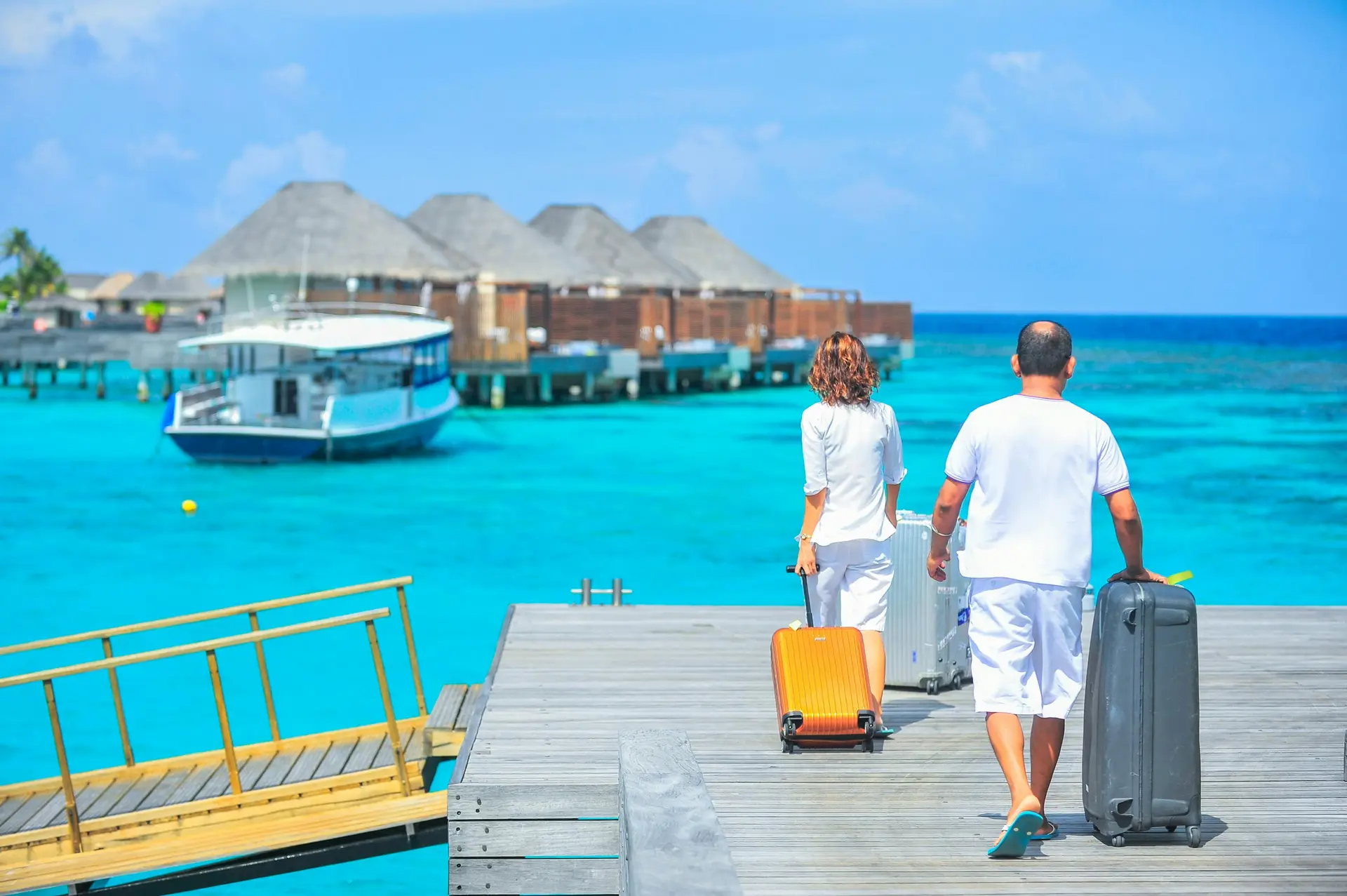 A man and woman with luggages walking on dock. There are small sea side villas can be seen in the distance.