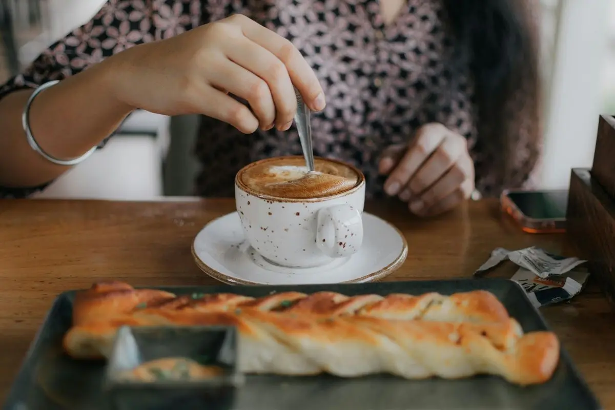 A person stirring their cup of espresso. Along with the cup, a bread can be seen on the table.