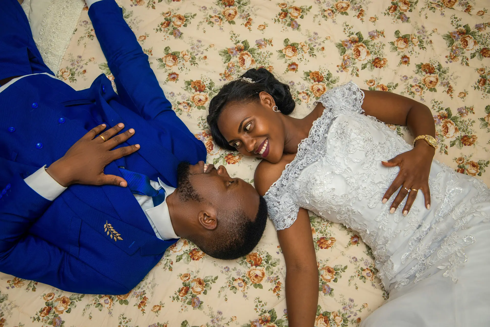 A man in blue jacket and a woman in white wedding gown laying on a bed.