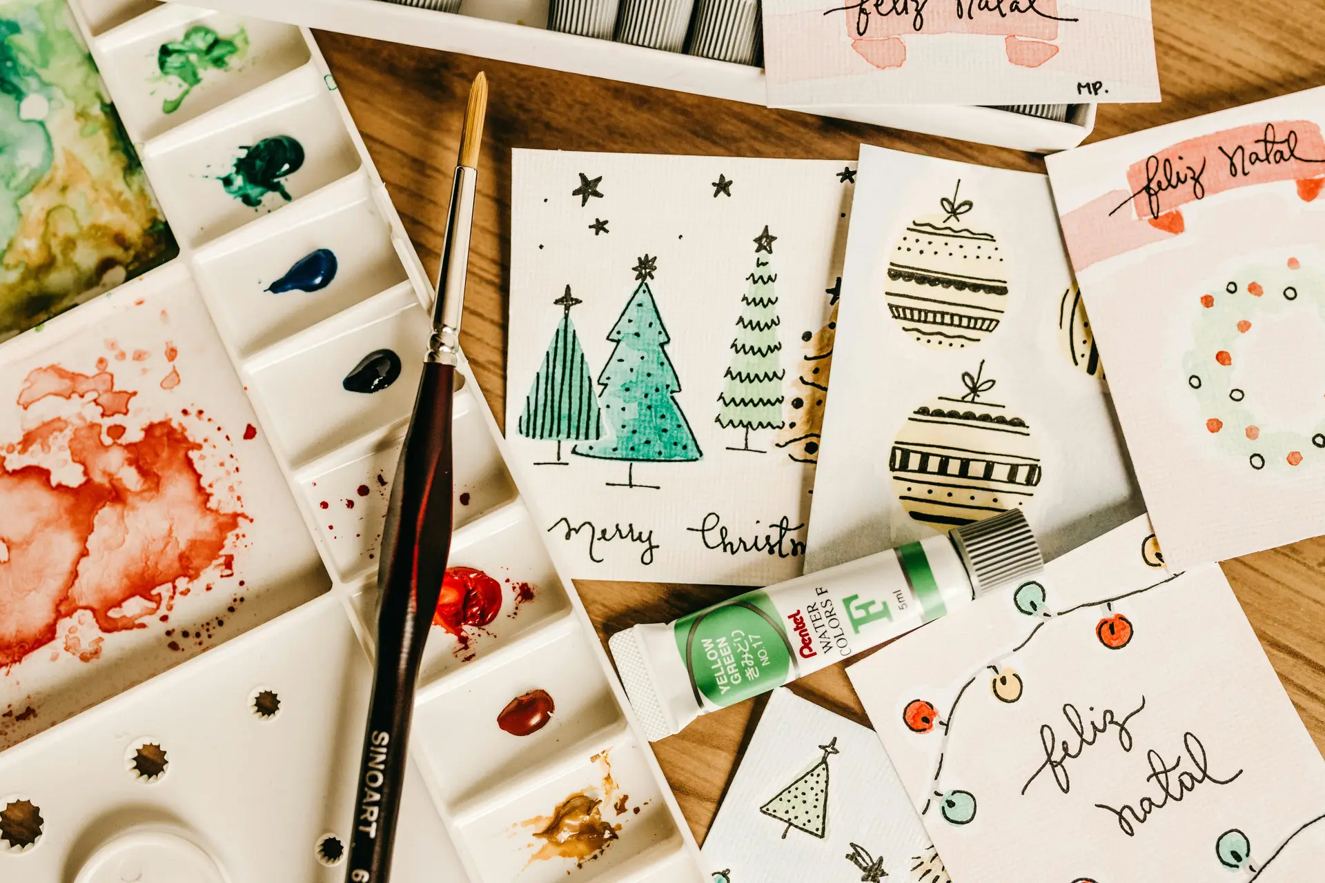 Paints, brush and other supplies on a surface along with handmade Christmas cards.