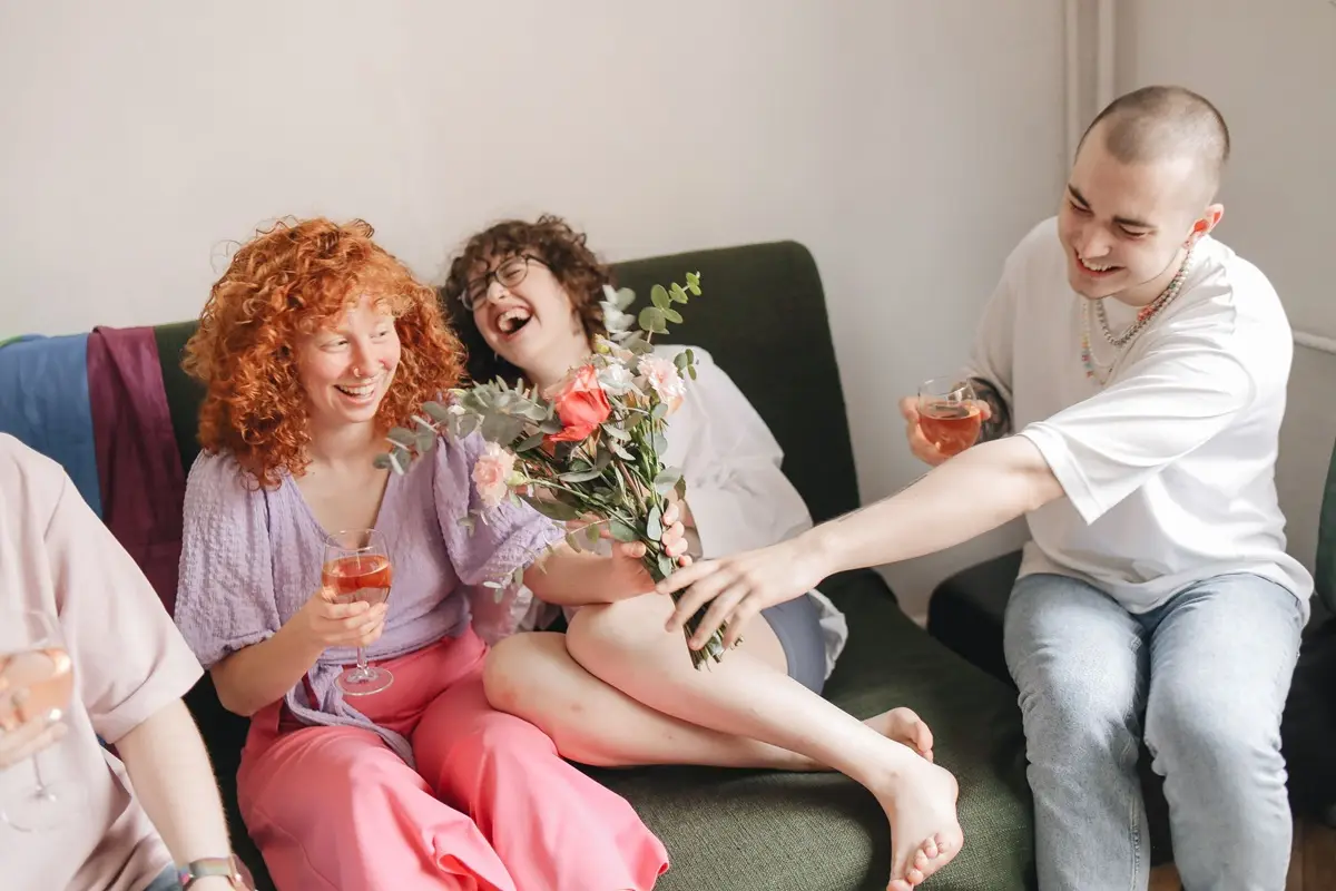 Group of friends laughing and enjoying wine on a sofa indoors with a bouquet.