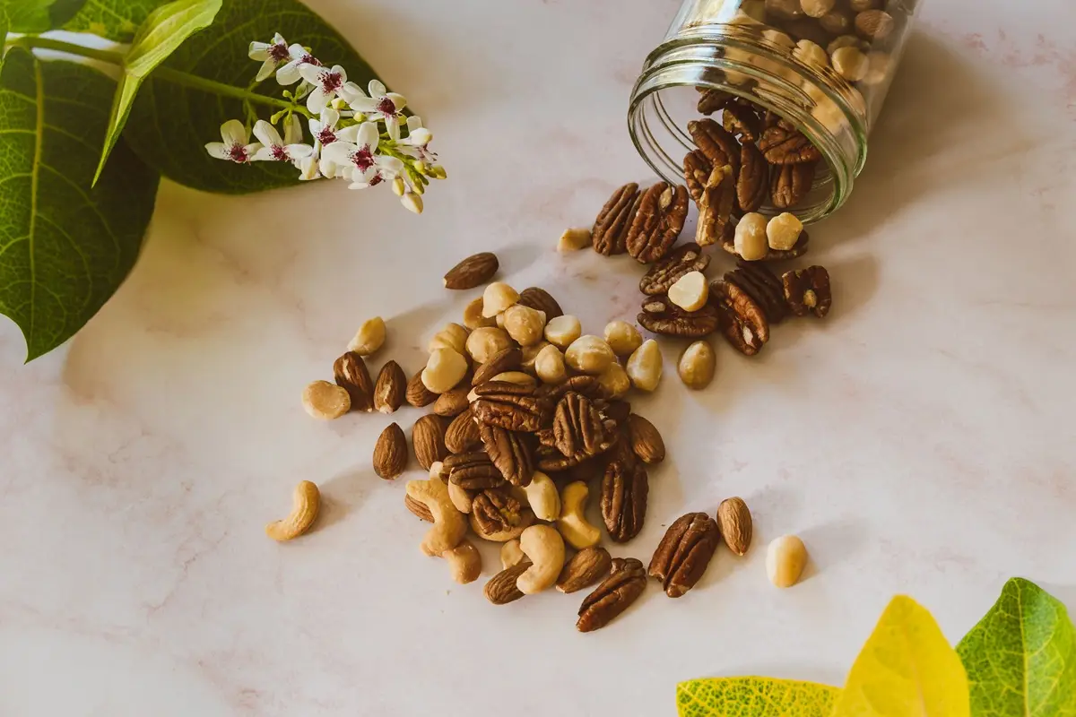 Assorted nuts spilling from a clear jar onto a marble surface with green leaves and white flowers.