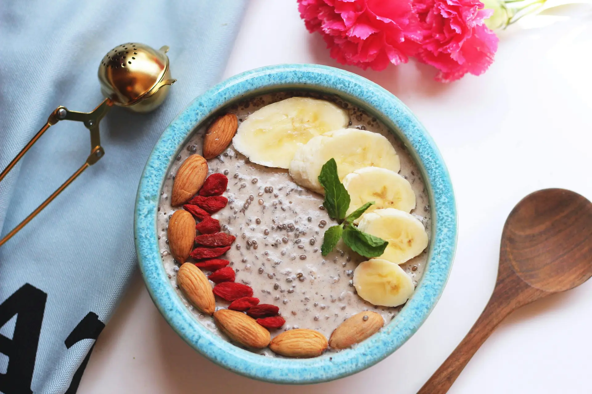 A bowl of nutritious food that has nuts and sliced bananas. There are wooden ladle, and flowers next to the bowl.