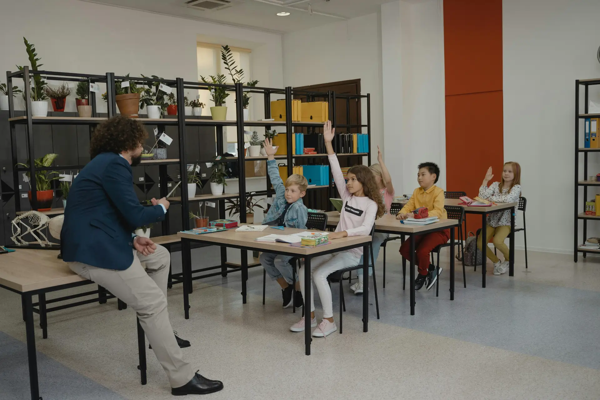 A teacher leaning on a desk and asking questions. Students have raised their arms from their desks.