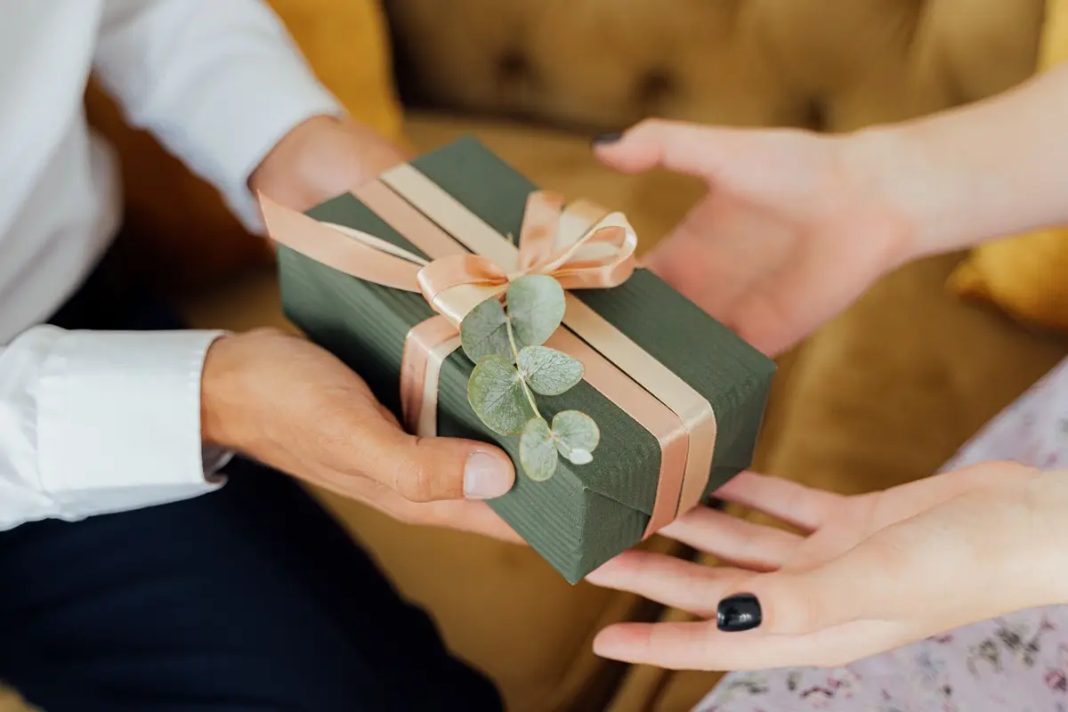 Two hands exchanging an elegantly wrapped green gift box with ribbons and foliage on sweetest day.