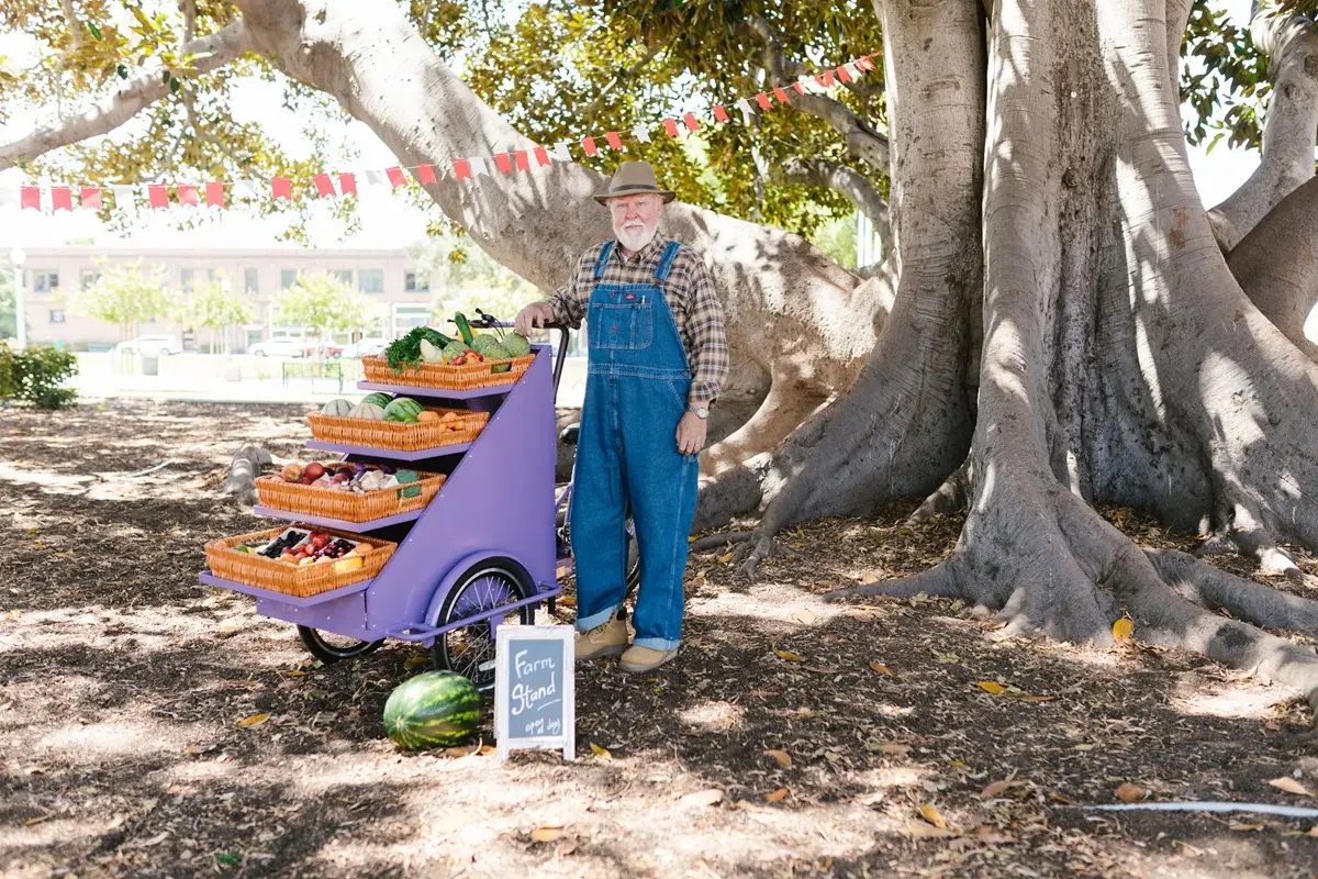 Senior farmer with a cart of fresh vegetables and fruits at a farmers market under a tree.