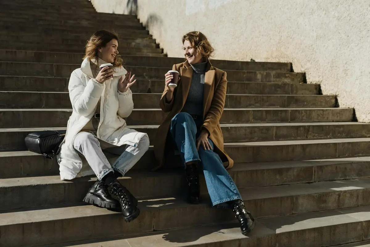 Two women sitting on stone steps enjoying warm drinks on a sunny day.