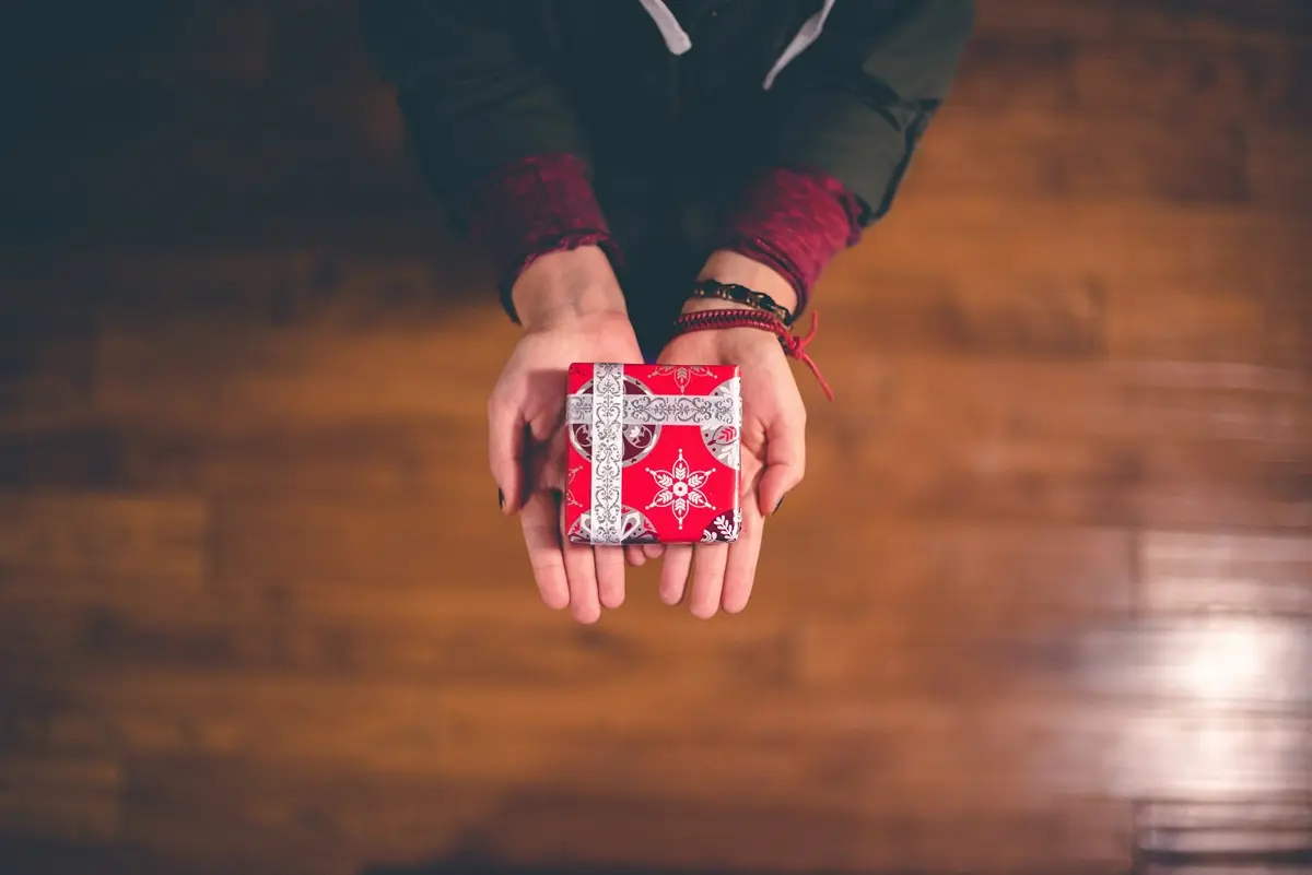 Two hands of a person holding a gift box wrapped in red wrapper.