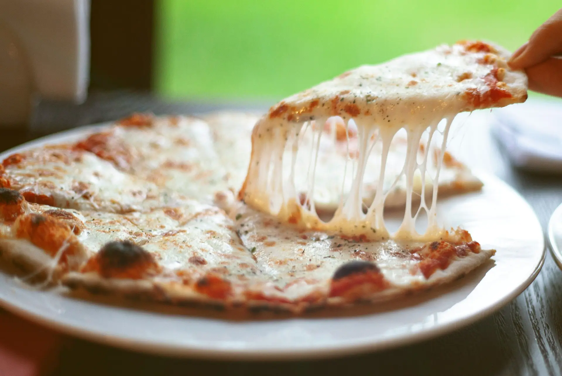 A person taking a slice of cheese pizza from a plate. The cheese is melted so much that its forming cheese strings.