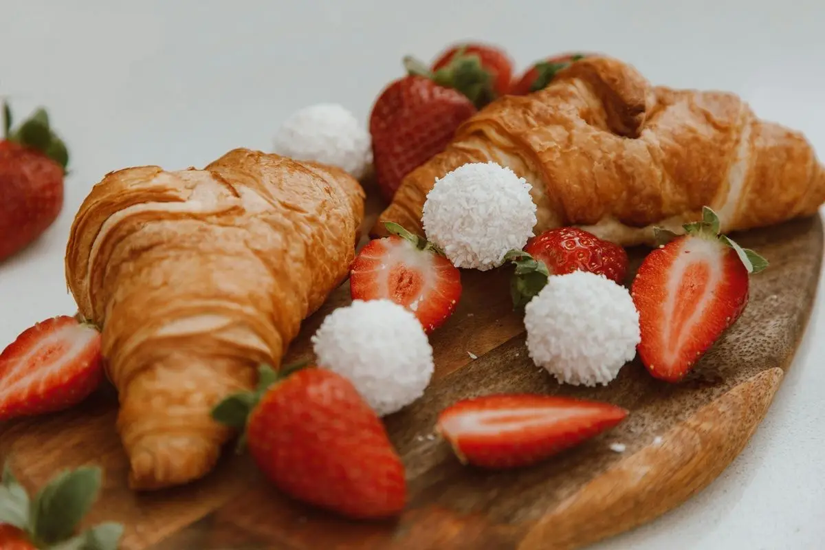 Two croissants on a wooden platter along with some fruits like Strawberry.