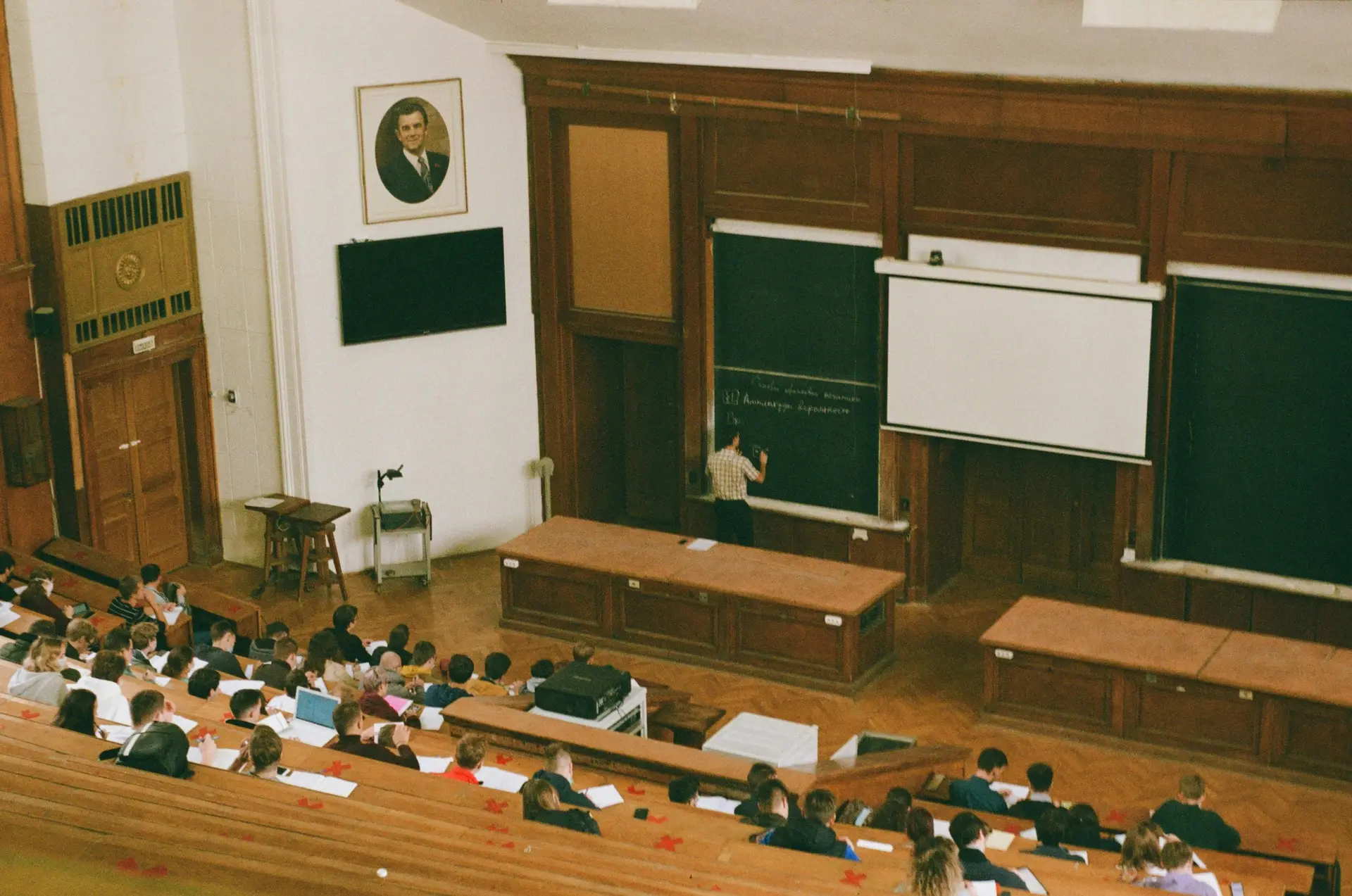 A teacher writing on a big black board in a classroom and the students are taking notes.