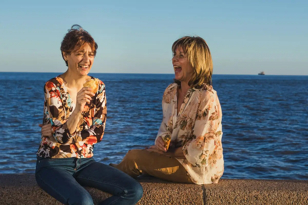 Two women laughing and eating by the sea in Montevideo, Uruguay, under a clear blue sky.