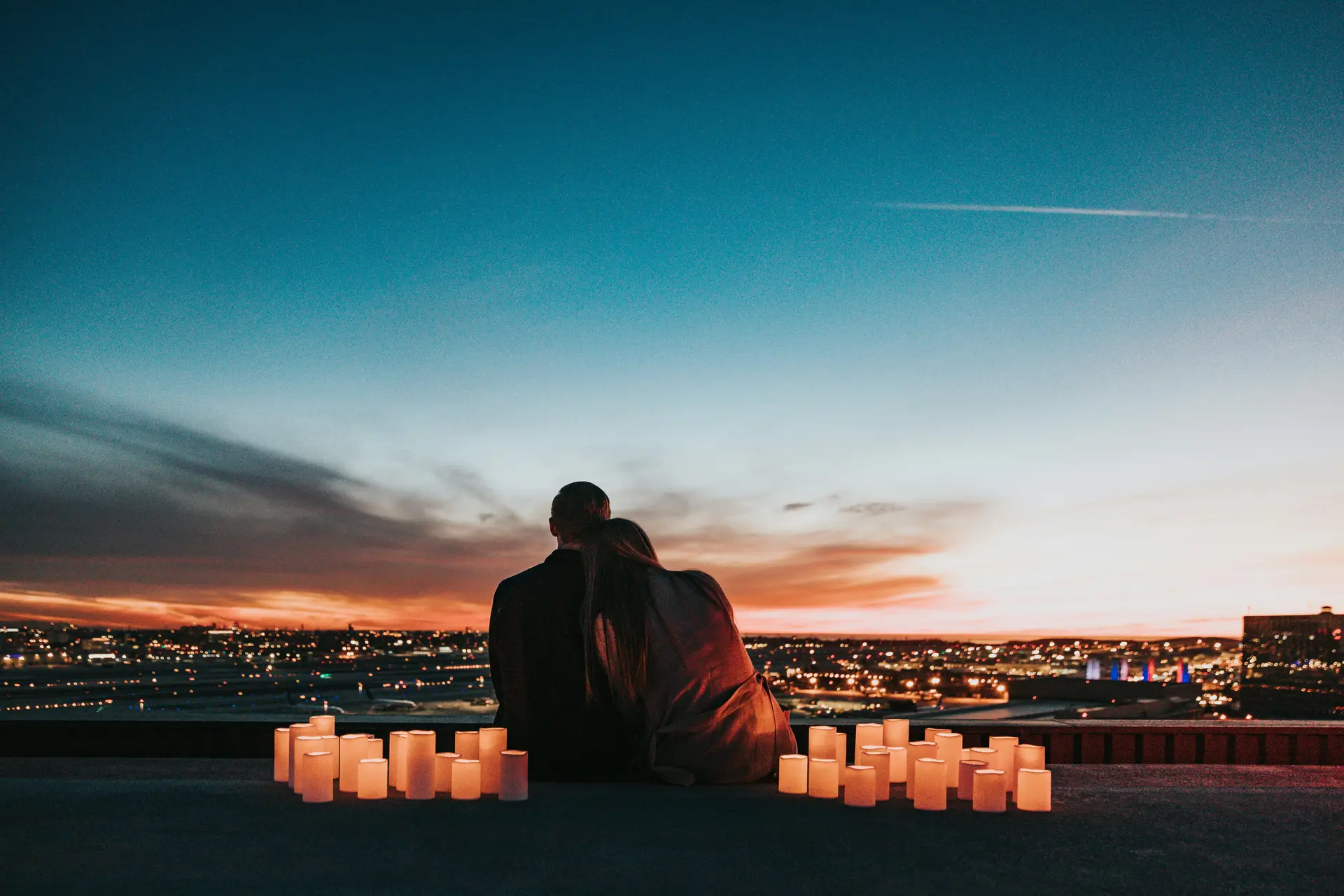 A couple sitting on the rooftop enjoying the sunset view. There are some already lit candles near them.