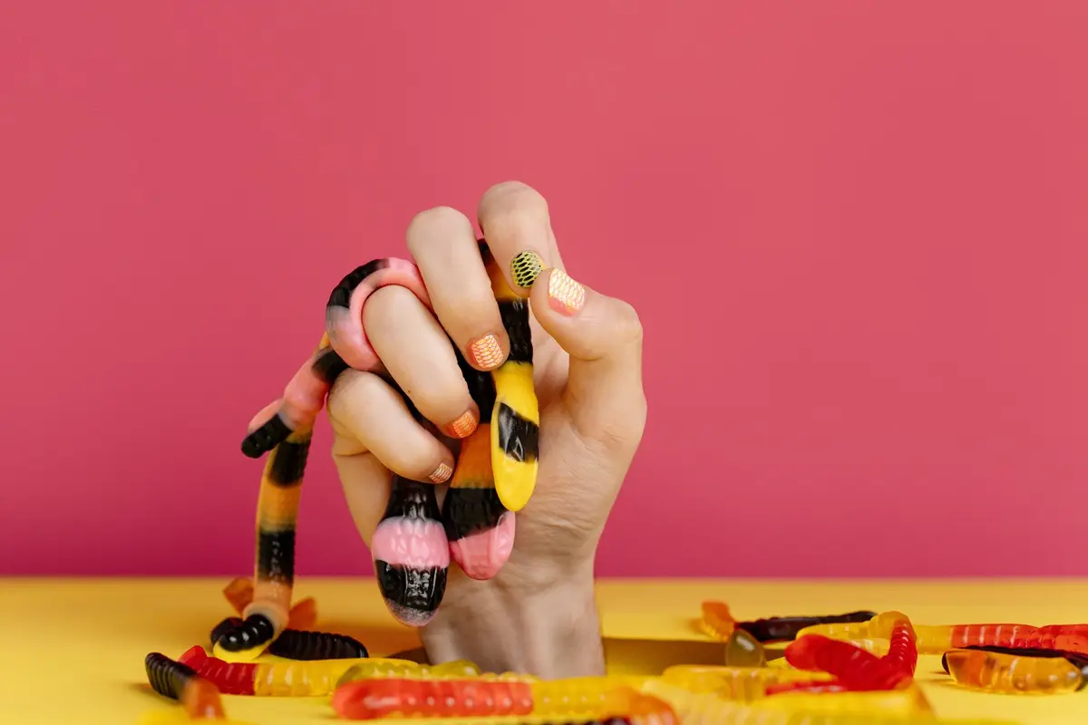 A hand with manicured nails gripping colorful gummy worms against a pink background.