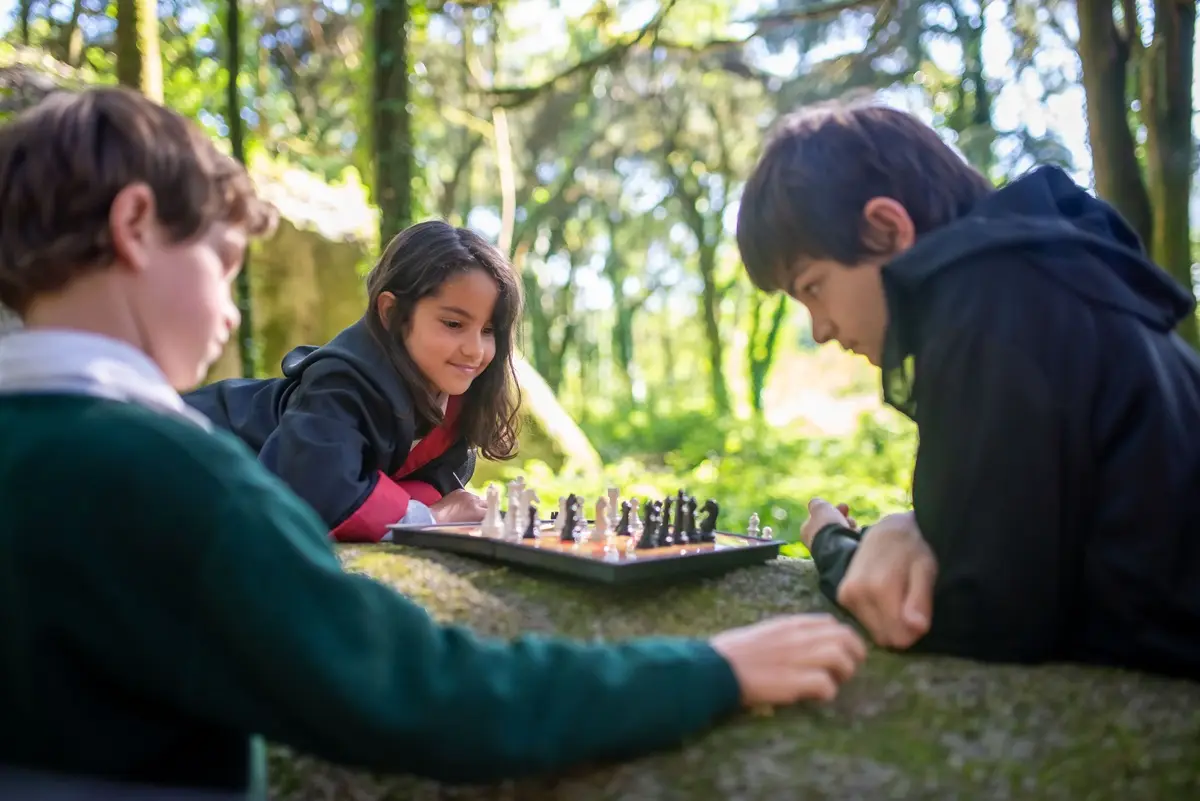 Three children in costumes playing chess on a stone outdoors in a forest.