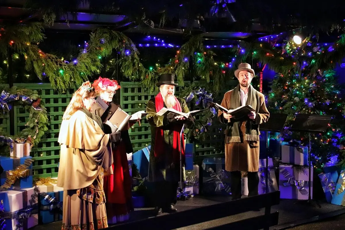 Four person wearing Christmas dresses are singing Christmas carols on a stage.